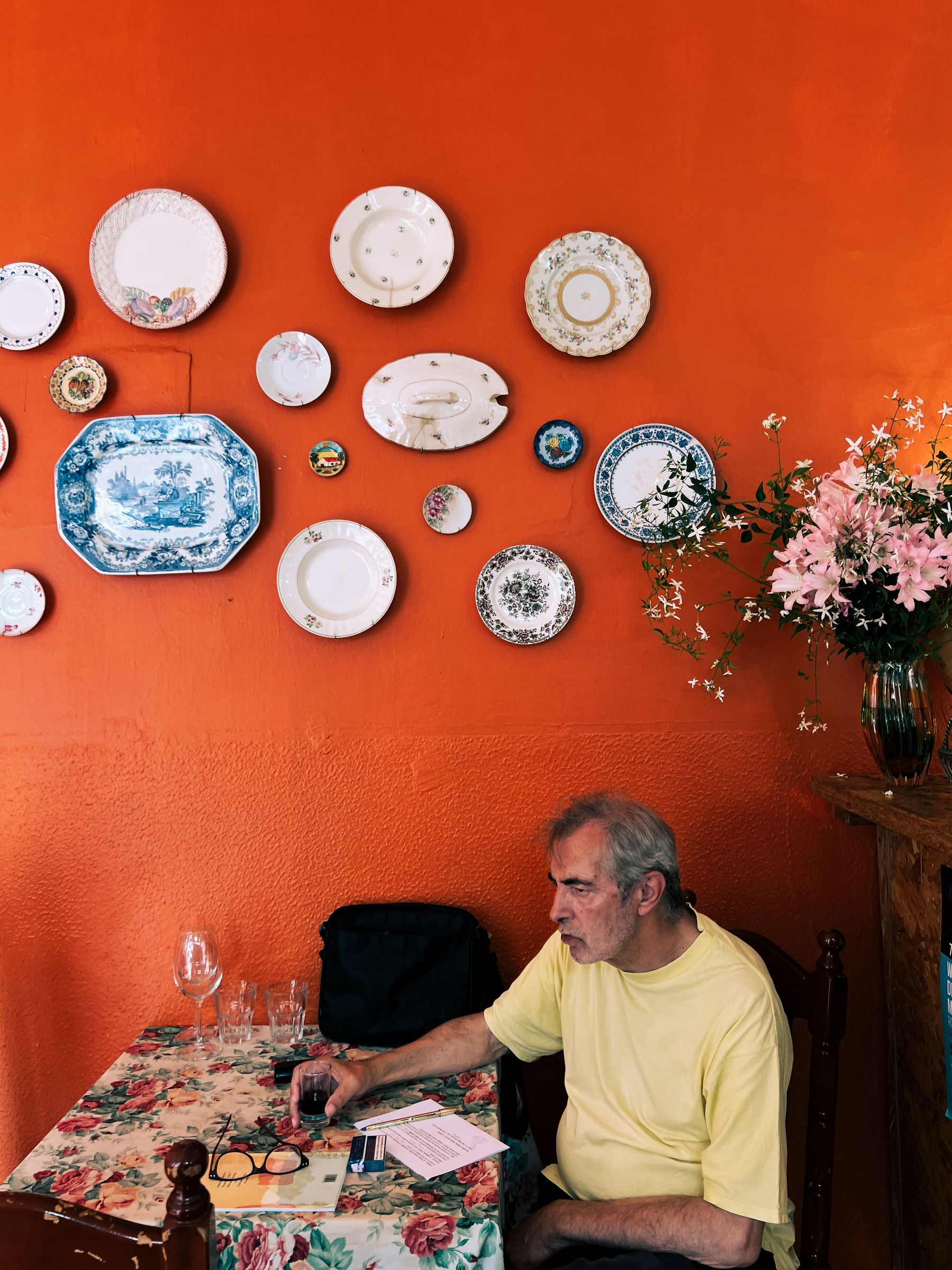 A man has a drink. Orange wall behind him, with hanging plates.