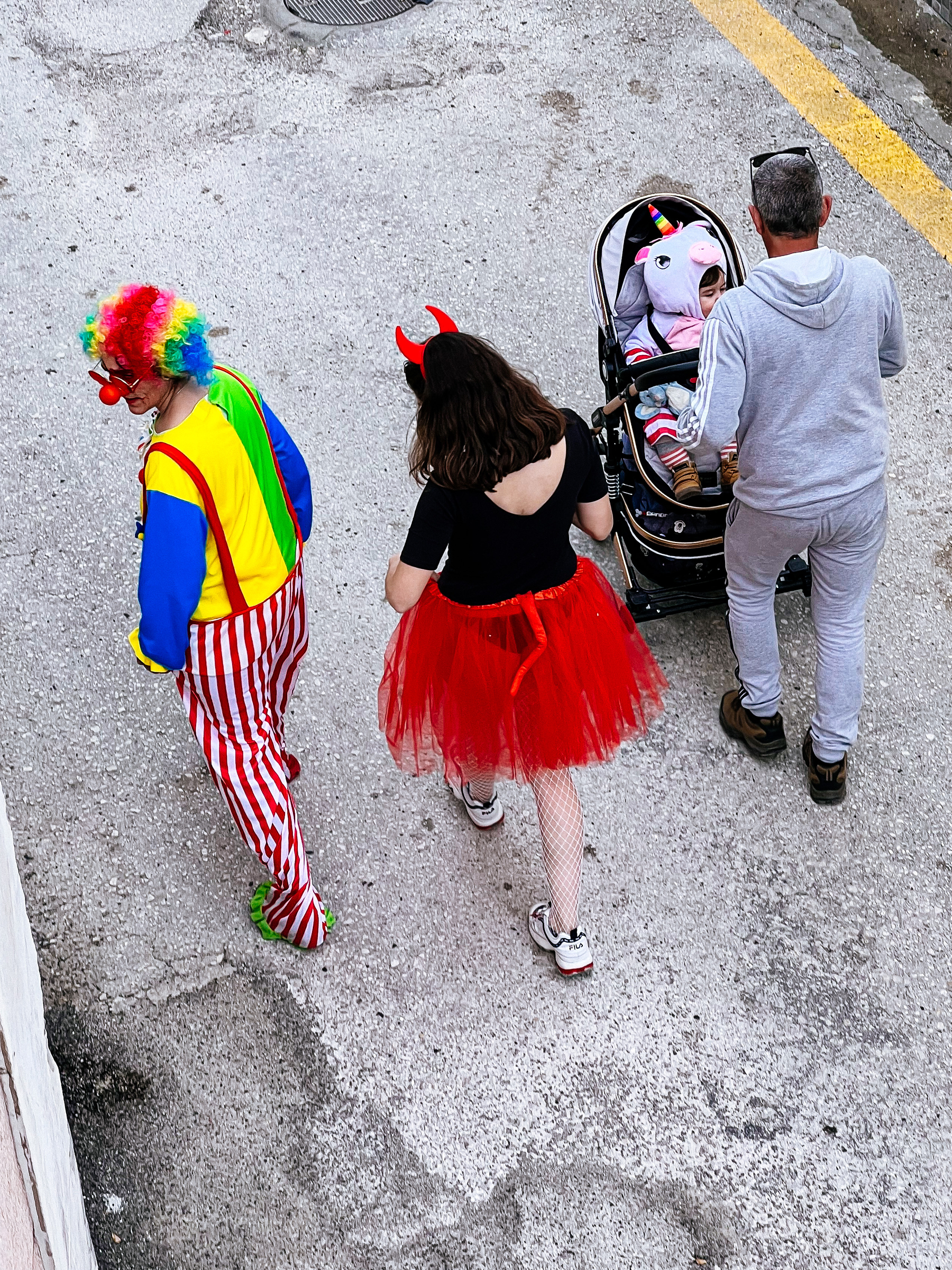 Three people walk away from the camera. We’re looking down on them. There’s a clown on the left, a woman with a red skirt and red devil ears in the middle, and a man with a grey track suit  pushing a stroller with a baby wearing a unicorn suit. Everything feels rather depressing. 