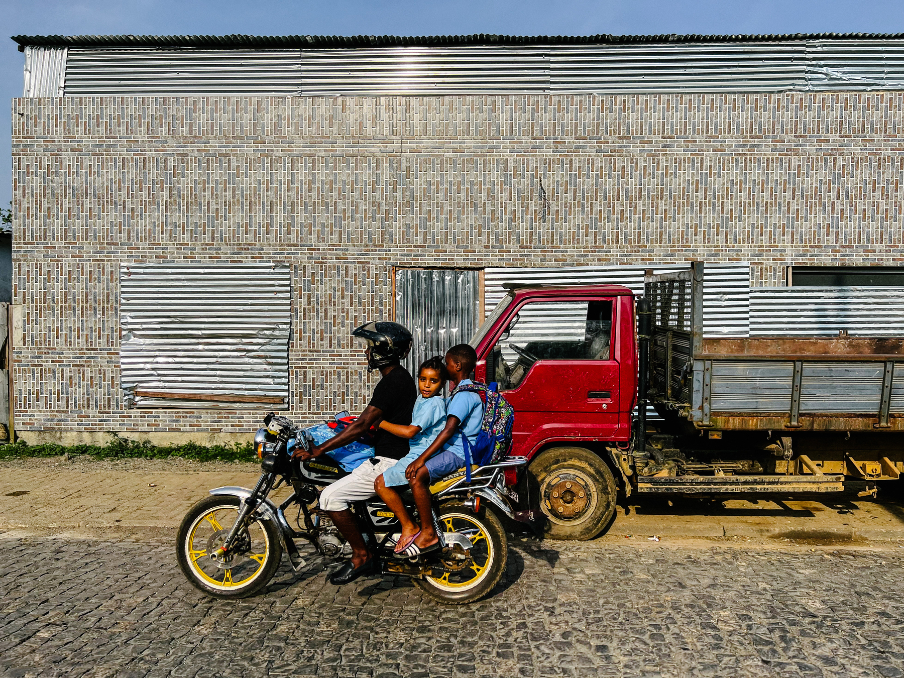 A father rides his motorcycle with his two kids behind him, in front of a very ugly building. 