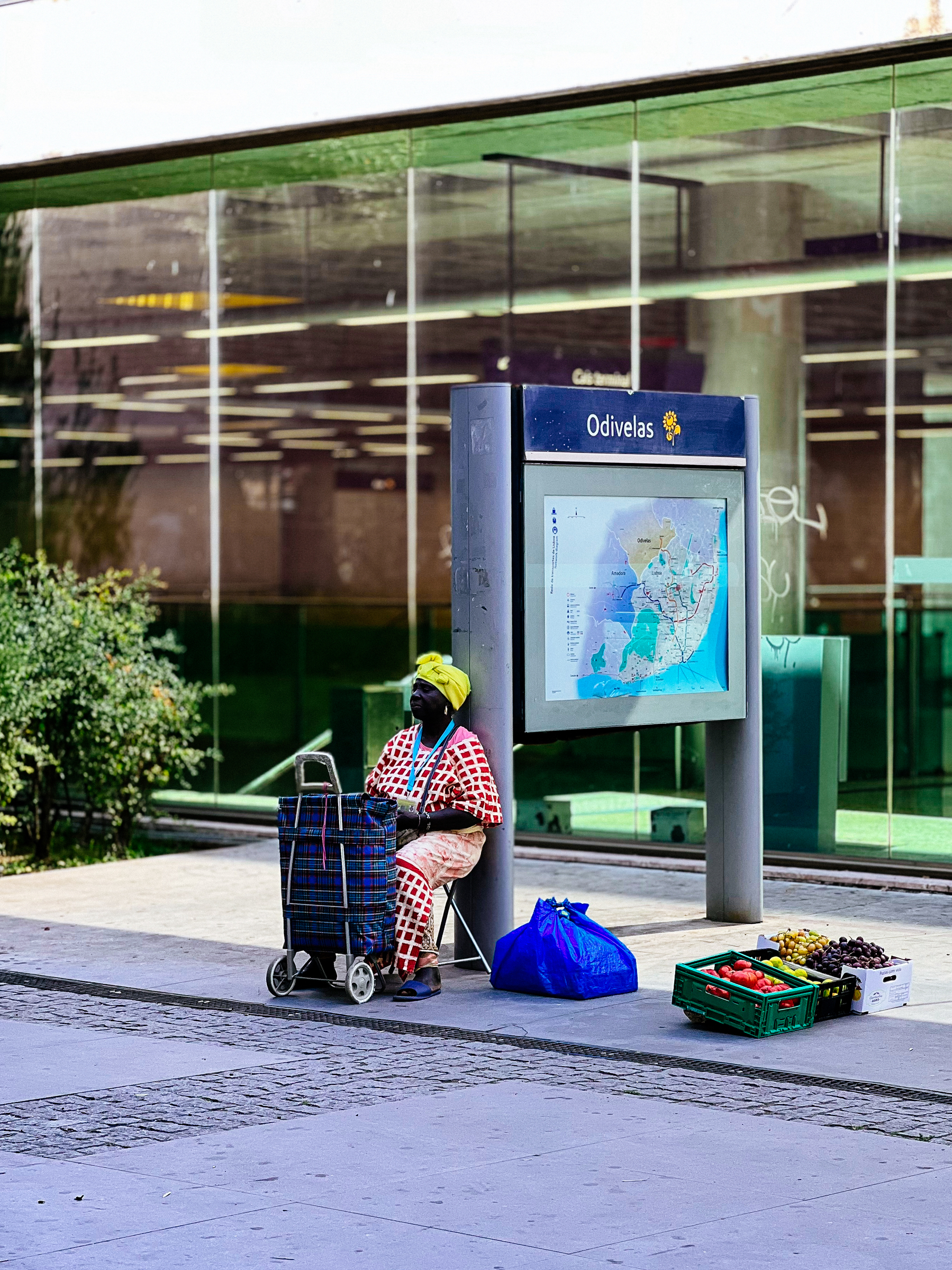A woman sit in a portable chair, selling fruits and vegetables by the metro station.
