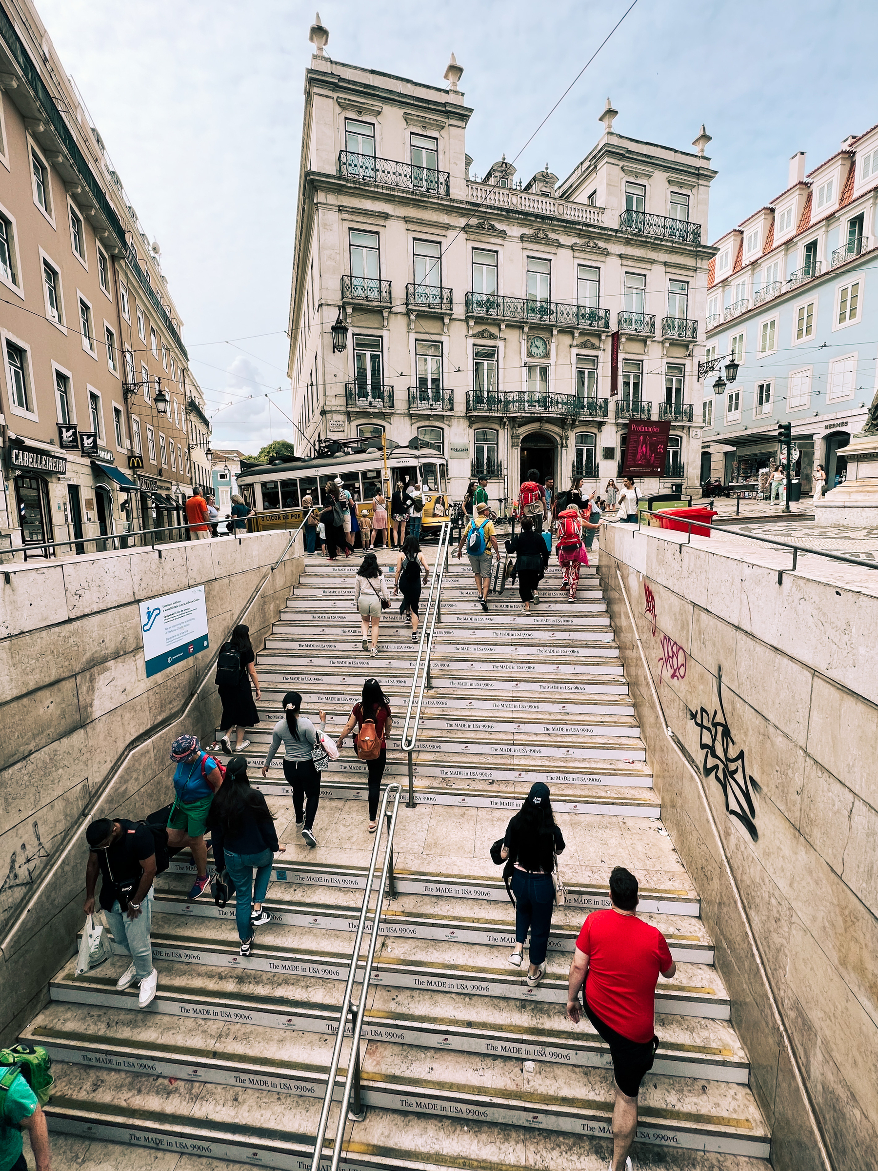 People on a stairway, coming up from a subway station, out into the center of town. Classic buildings, a tram.