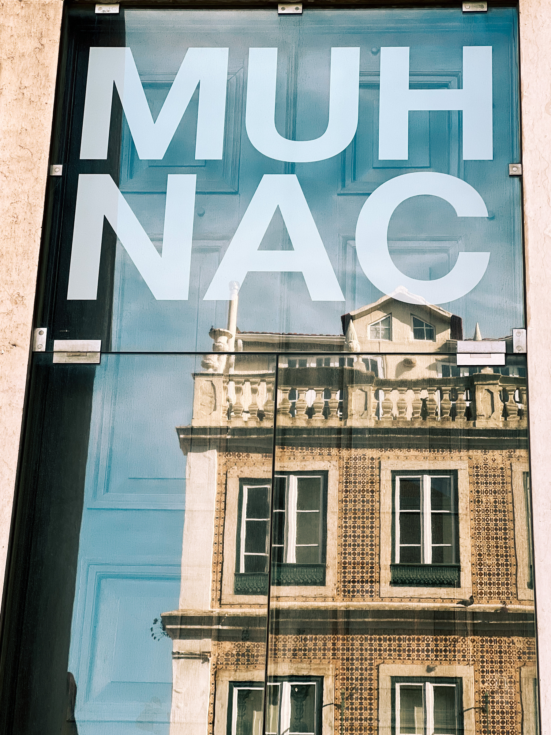 An old building reflected on a glass door, with MUHNAC written on it. 