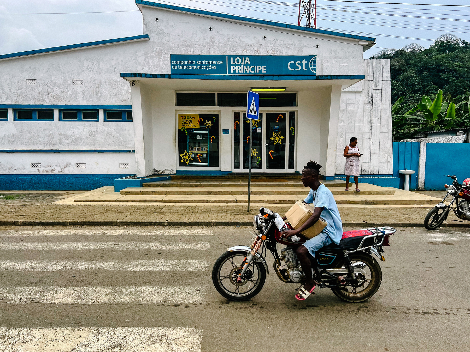 A motorcycle rides past the telecom office, a retro white building, with some blue thrown in. 