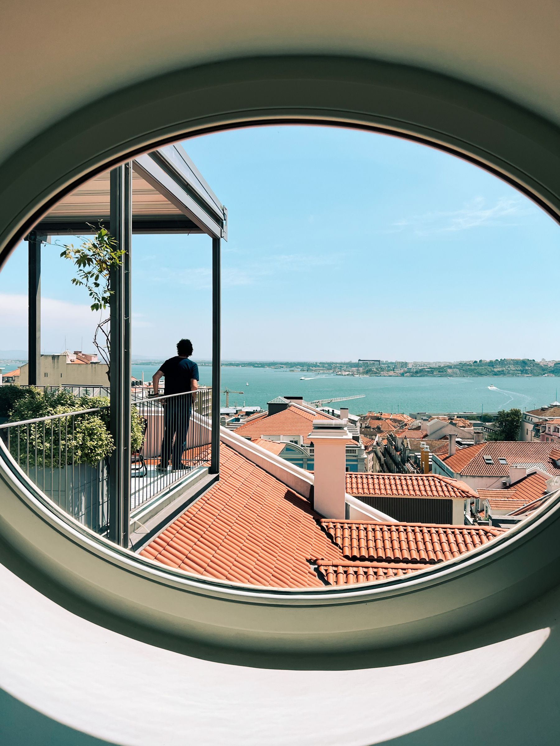 A man looks out into a sea of rooftops, and the river beyond that. He’s standing on a balcony, and we’re looking at him from inside the building, through a round window. 