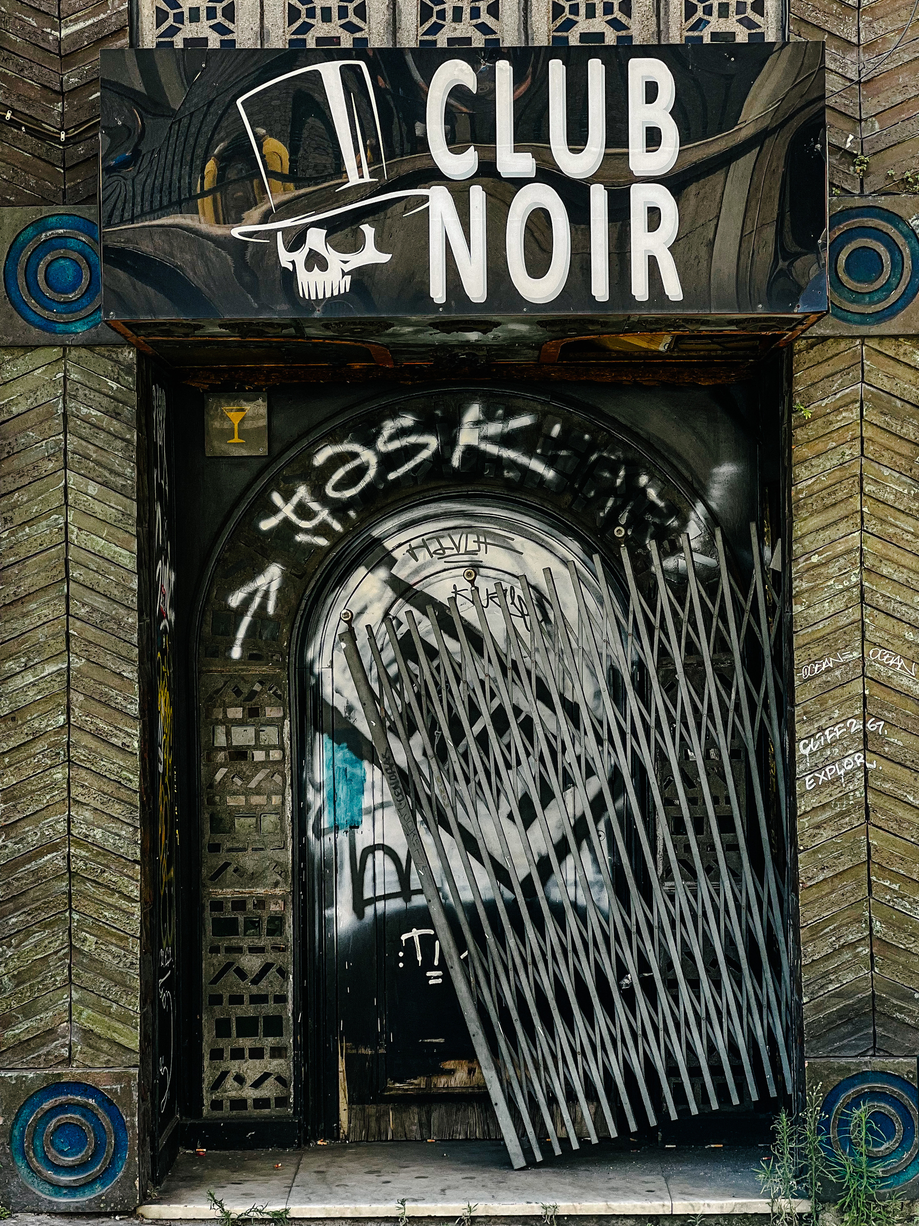 A run down club, “Club Noir”, probably closed for many years. There’s a broken gate, and a skull wearing a top hat. 