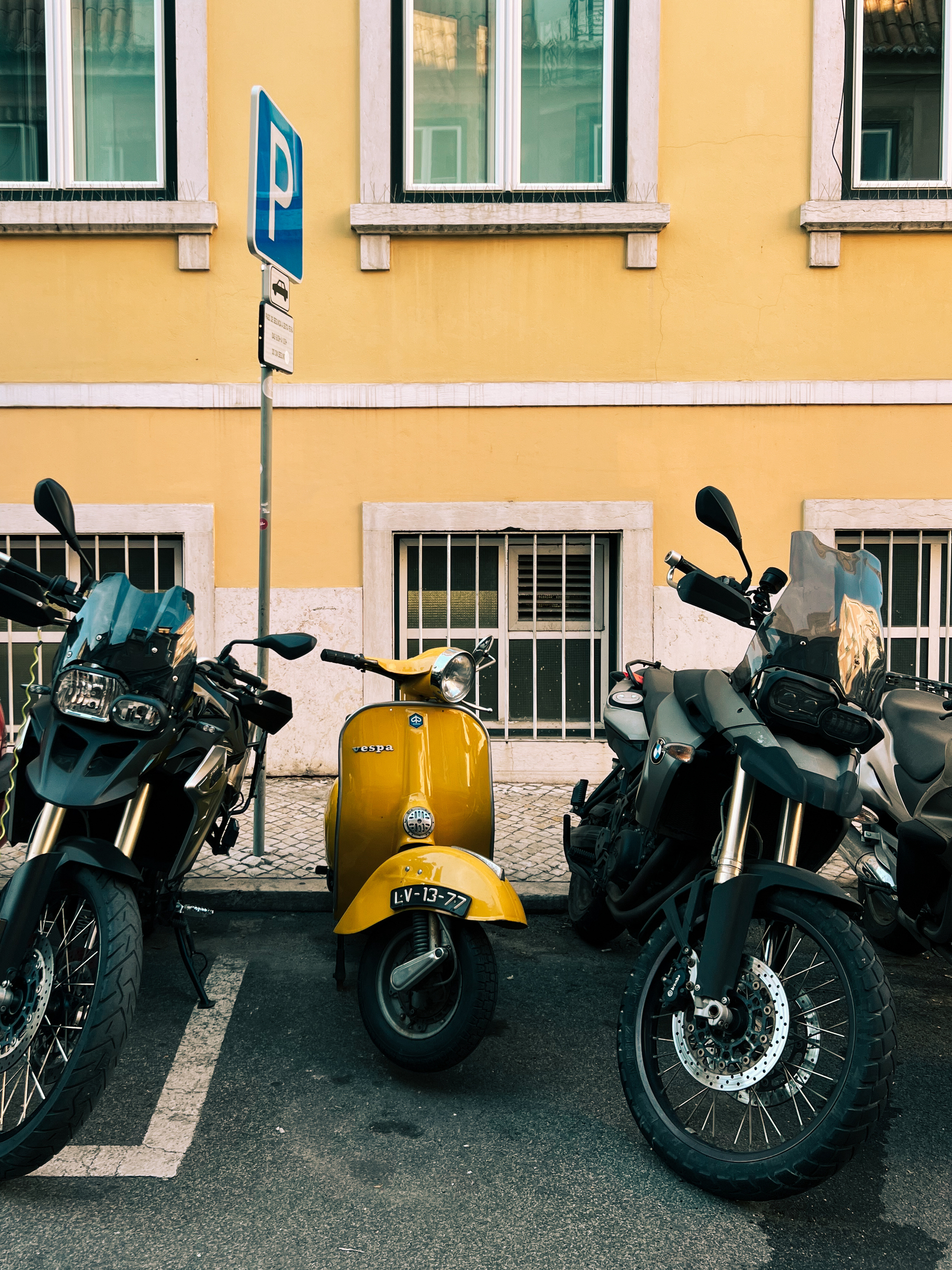 A yellow Vespa, with modern company, by a yellow building. 