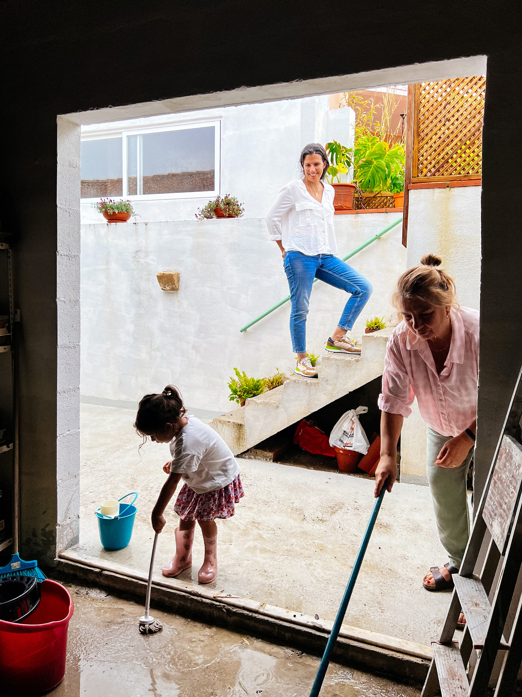 A toddler and a woman mopping, while a second woman looks on from a staircase in the back. 