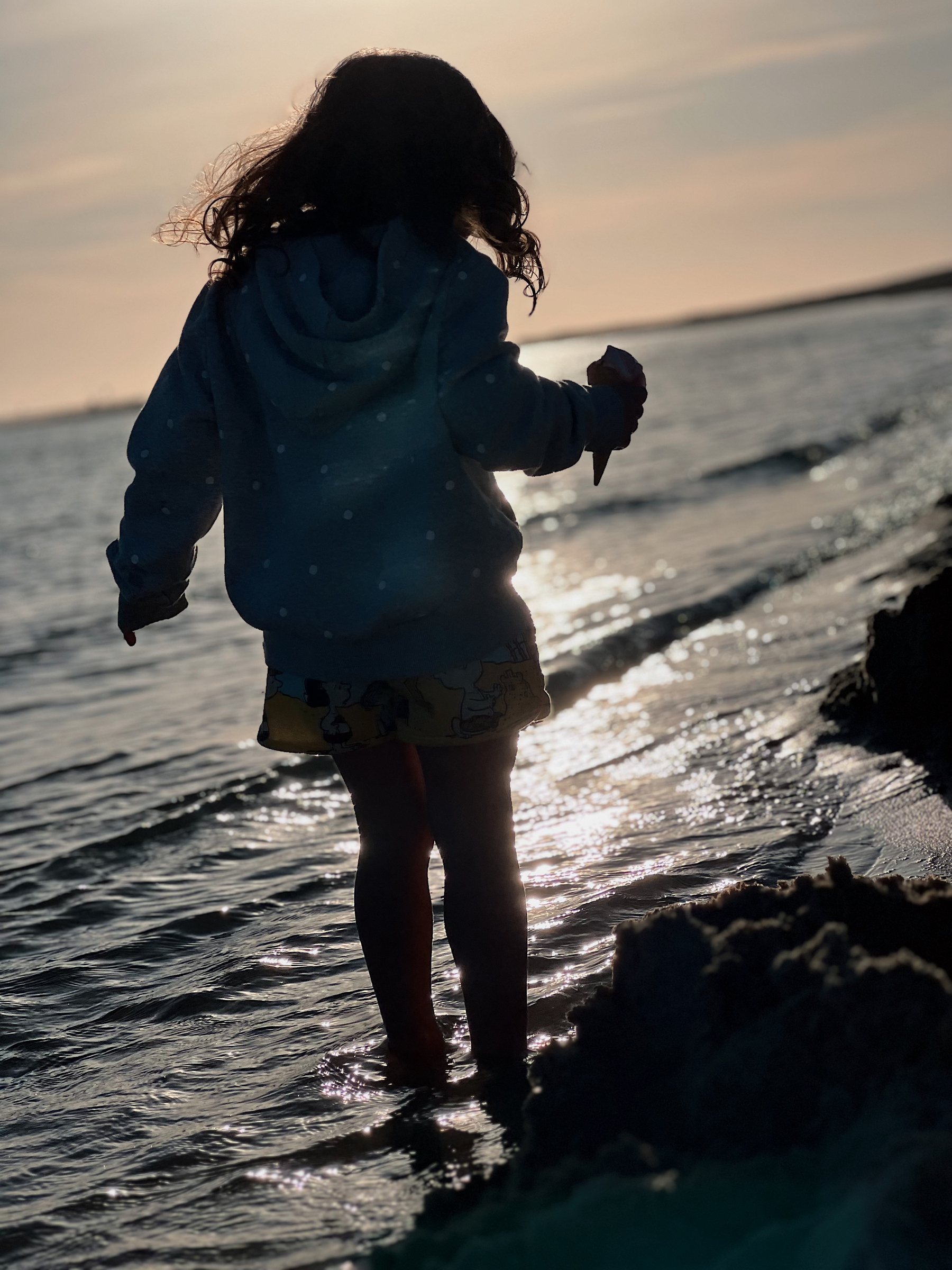 A girl walks along the shore, ice cream in hand. 