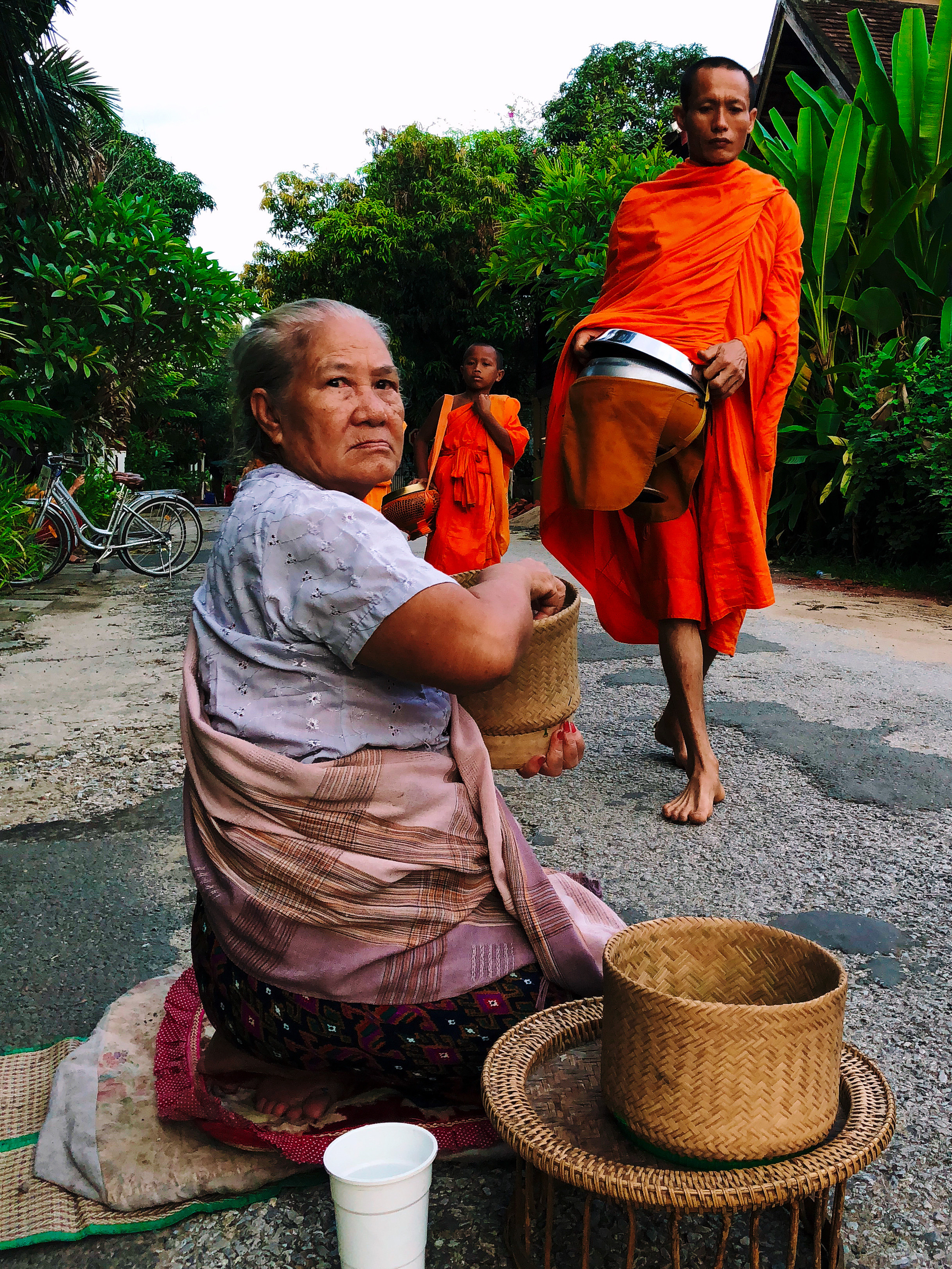 An old woman looks on, as monks walk by during Alms, wearing their orange robes
