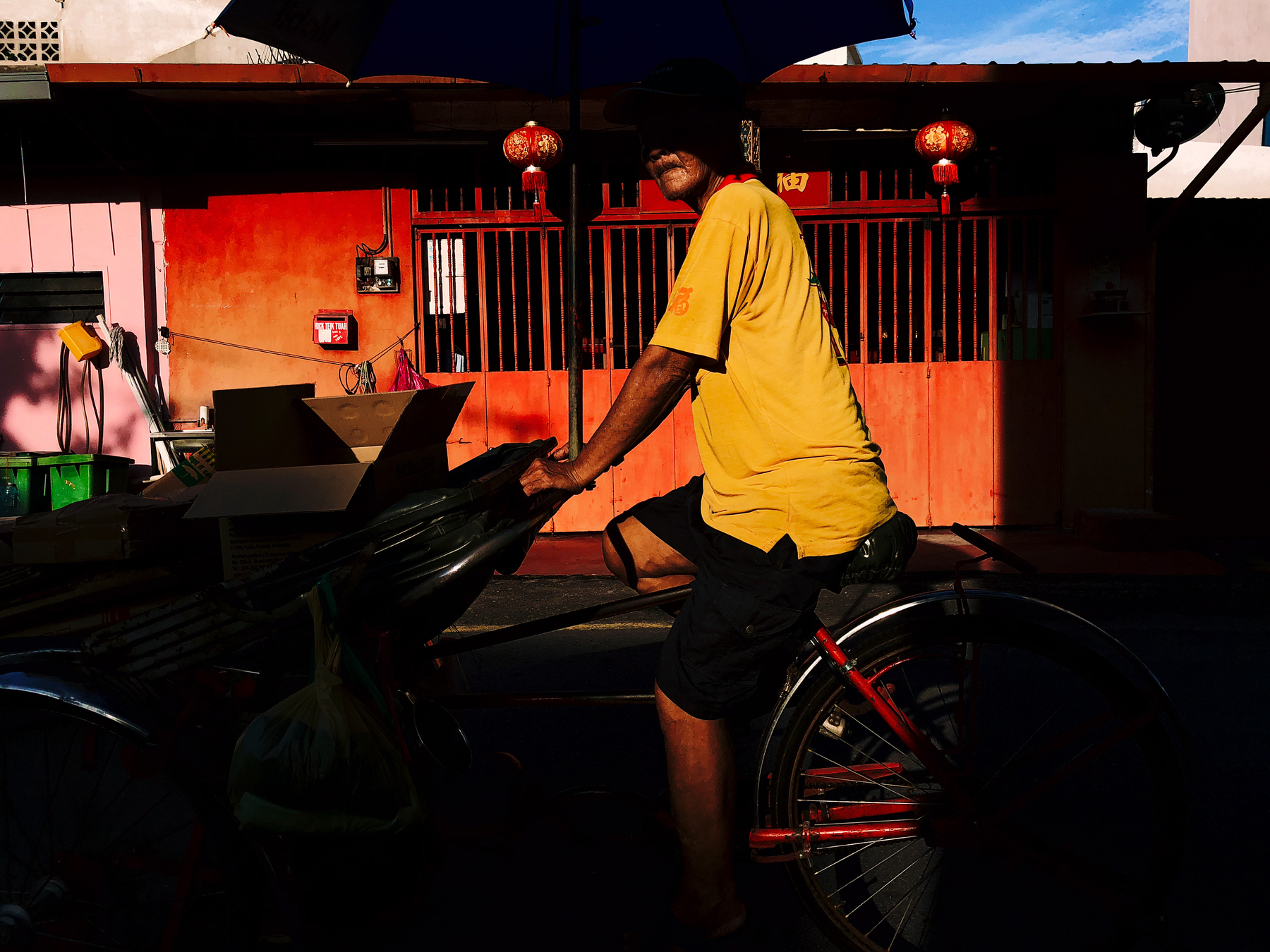 A man slowly cycles in front of us, in a very colorful scene. Yellow shirt, red background, an umbrella casting shade over his face. 