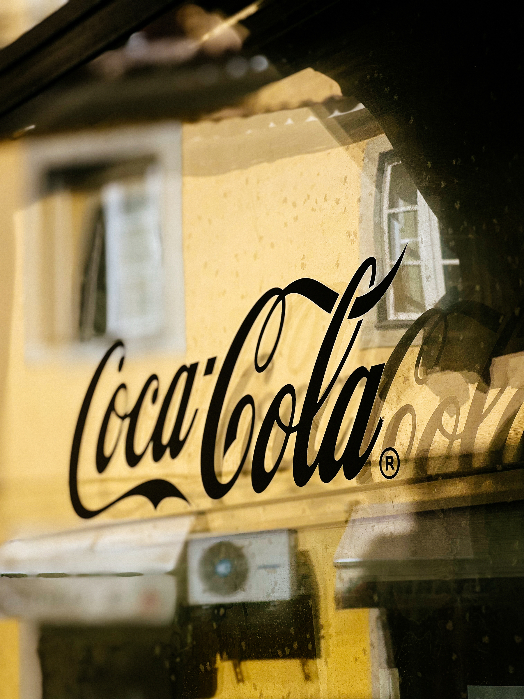Coca-Cola on a window, with a yellow building in the back. 
