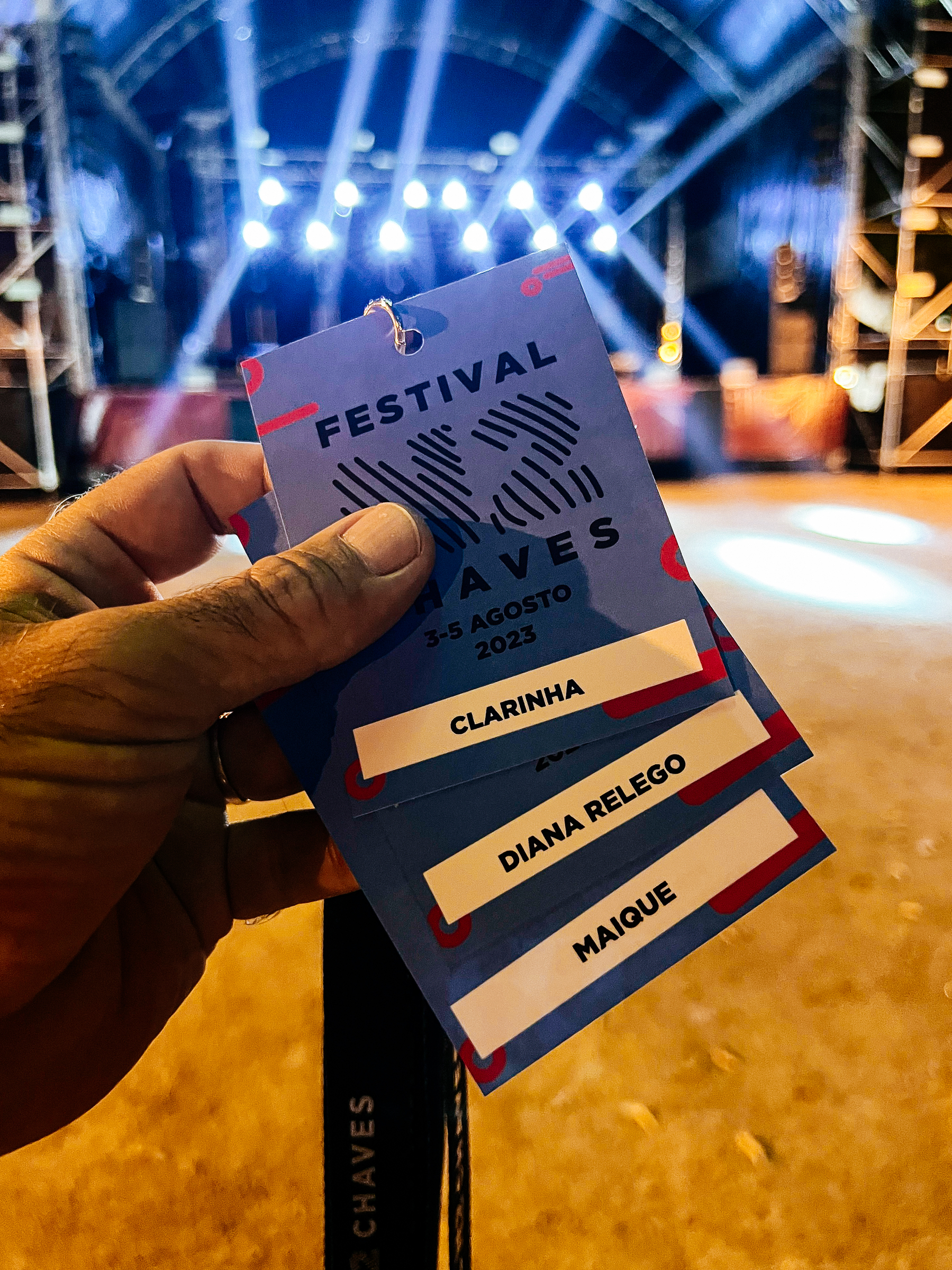 A hand holds three passes for a music festival. One for tinyMovieStar, one for MovieStar, and another one for me. There’s a stage with lights in the background. 