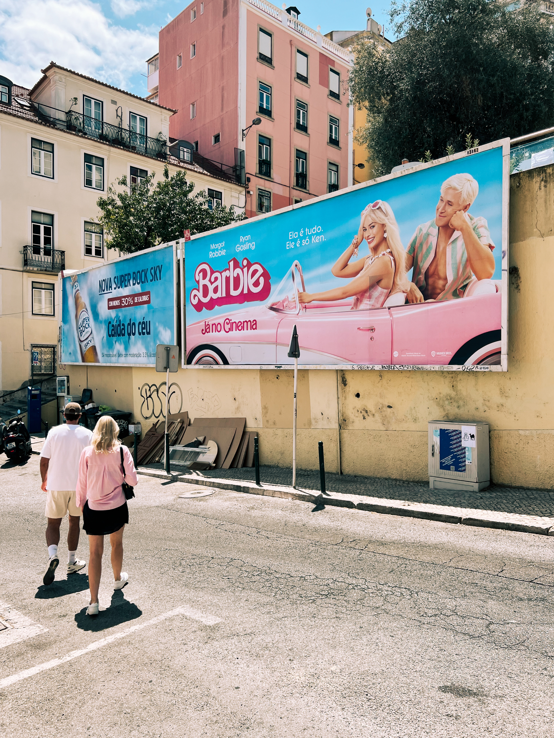 A Barbie movie poster, with a pink building behind it, and a girl with a pink shirt walking by. 
