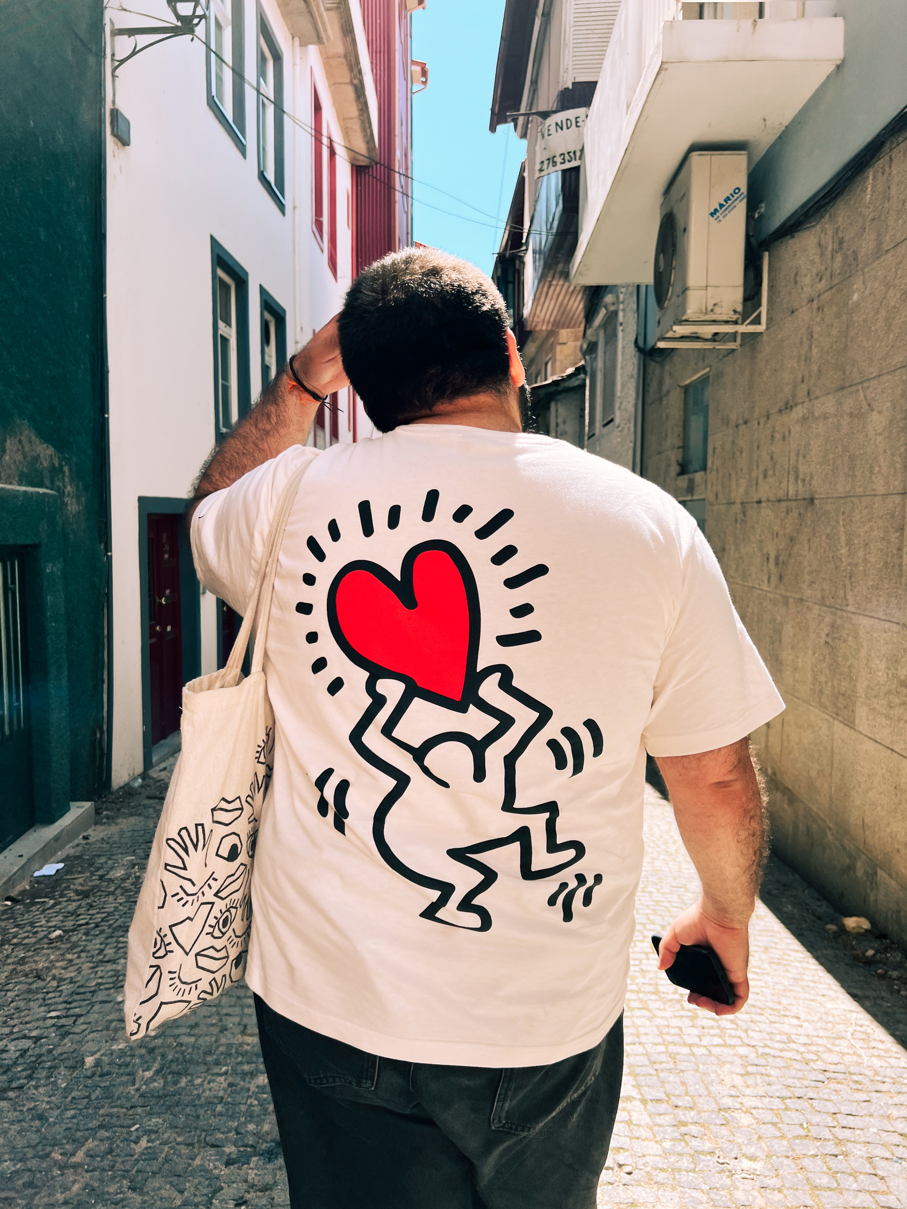 A man walks away from us, wearing a Keith Haring t-shirt, with a man holding a heart. 