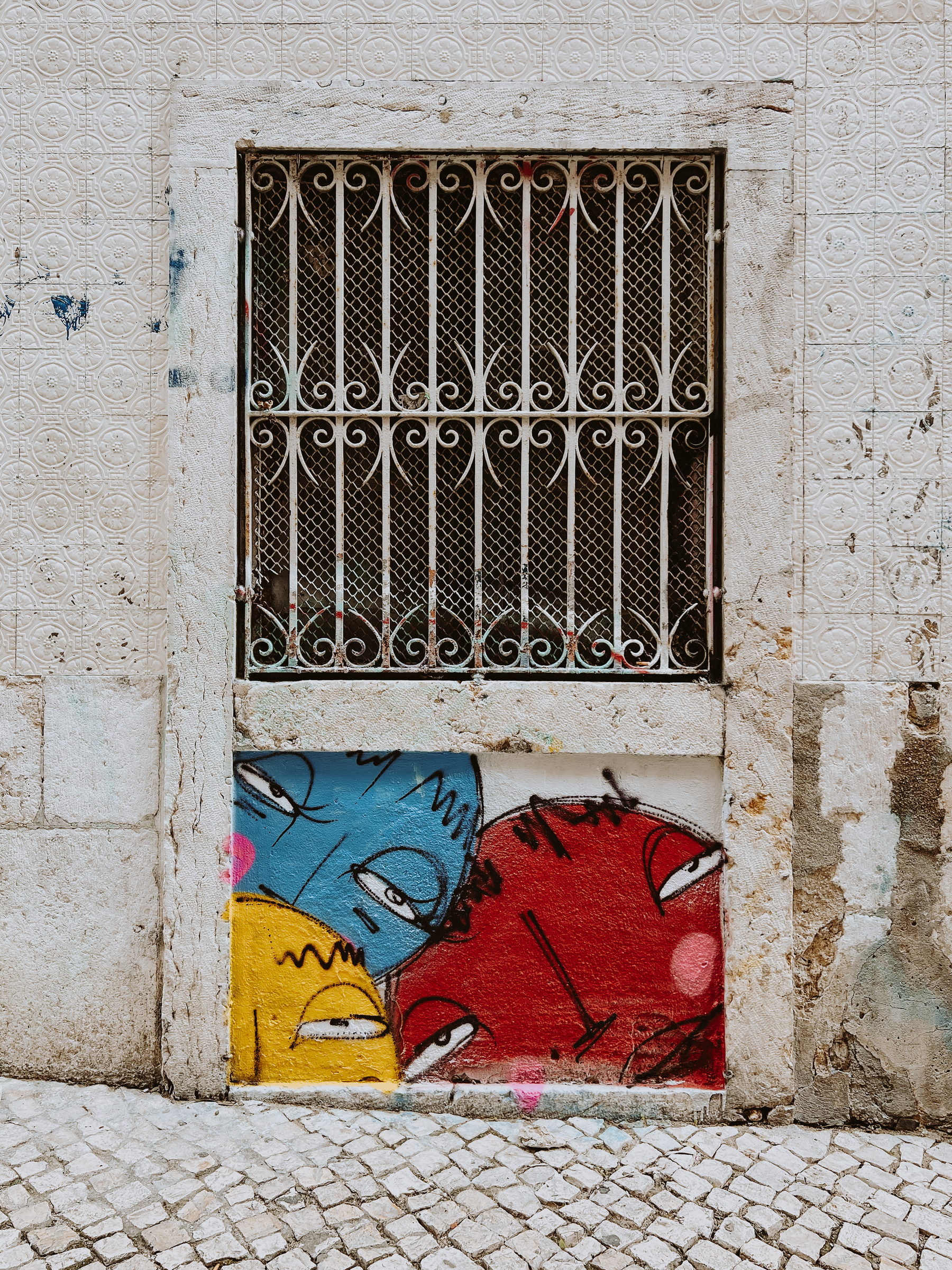 Street art. Three cartoony faces are graffitied under a window. The window has bars.