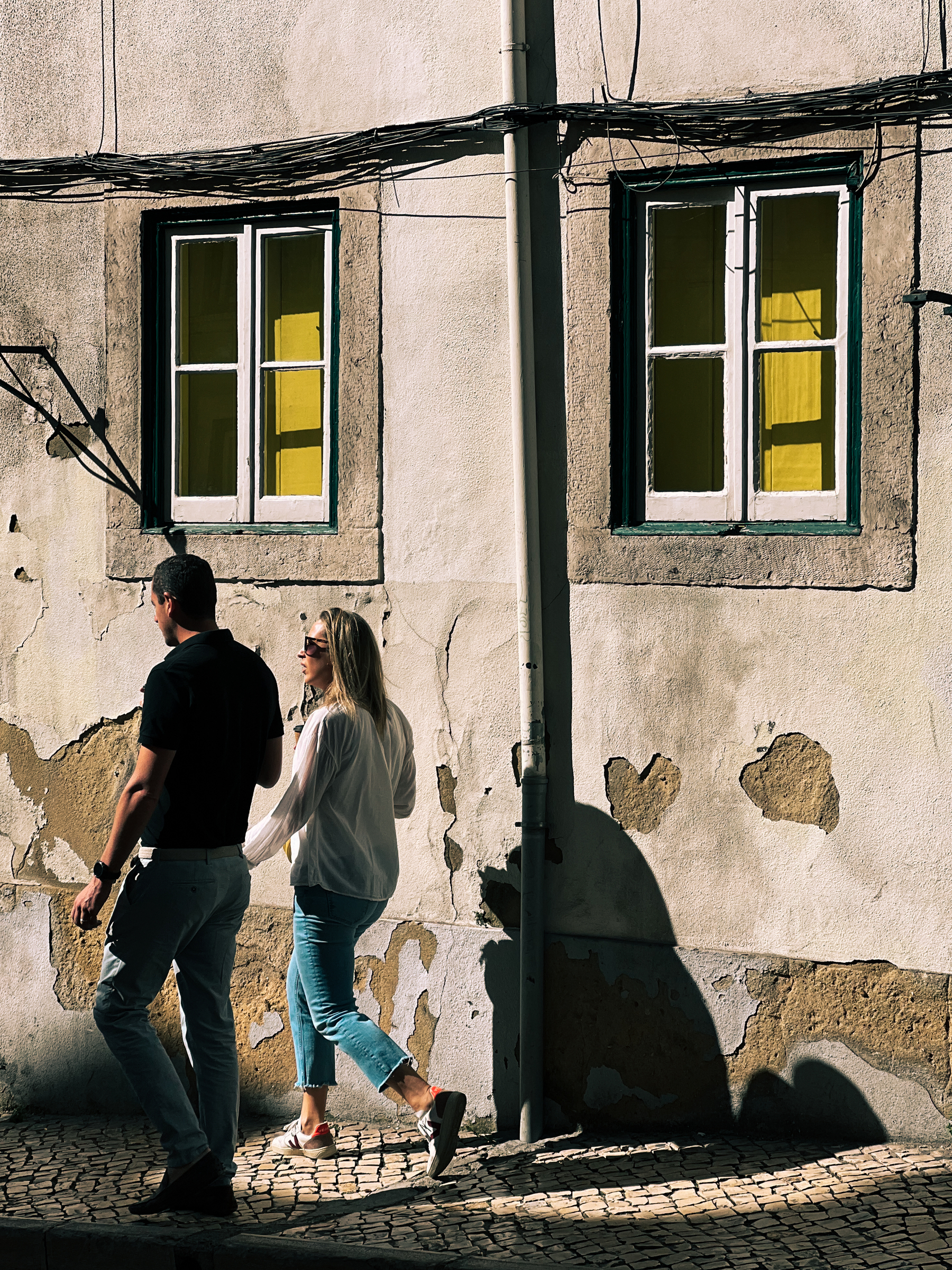 A couple walks by a poorly kept wall. Yellow windows. 