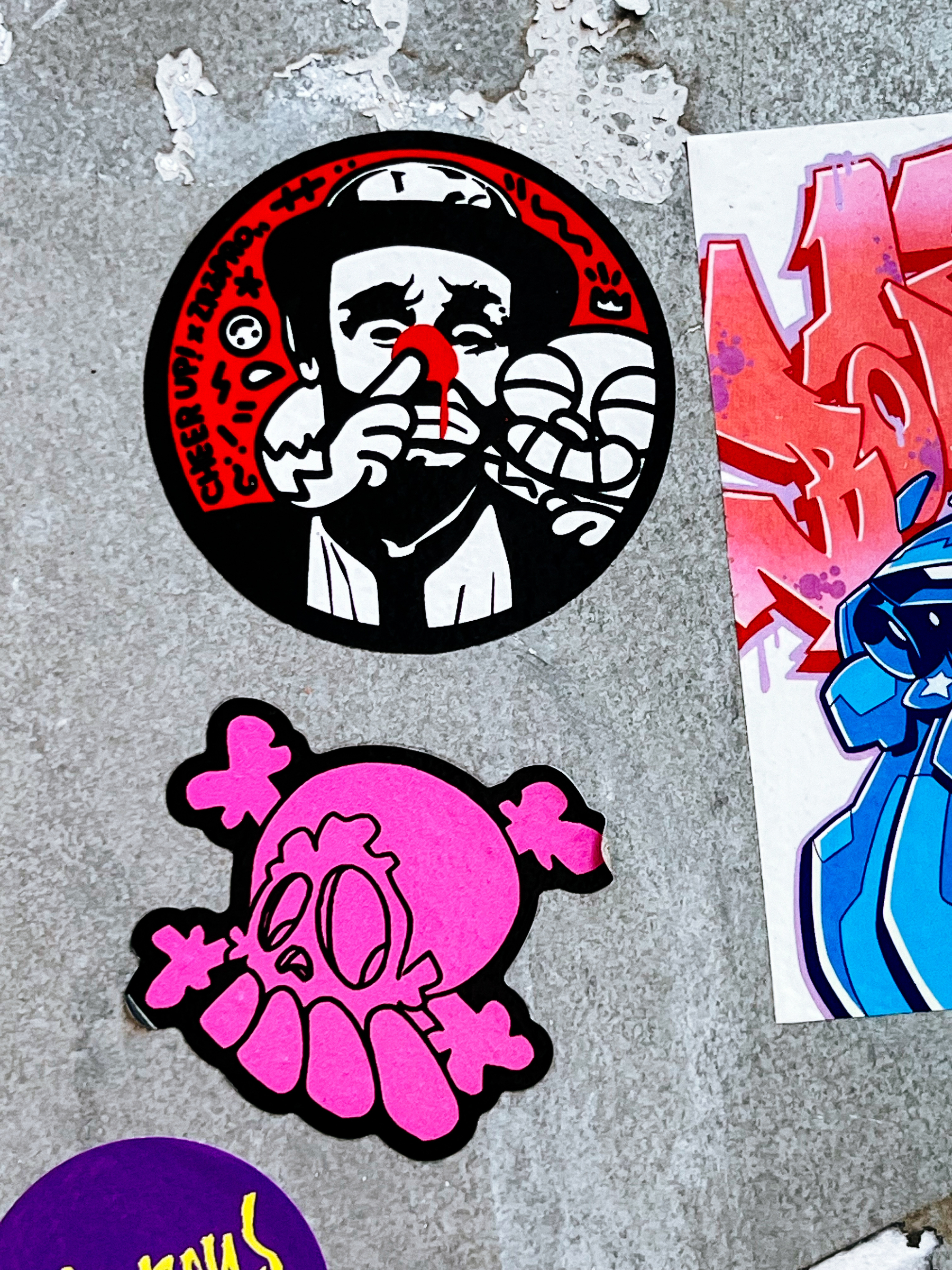 A two sticker combo. One has a robot like creature sticking a finger up a clown&rsquo;s red nose (yup, true), and the other one is a pink skull &amp; bones, with what looks like overdeveloped teeth (yup, true again).