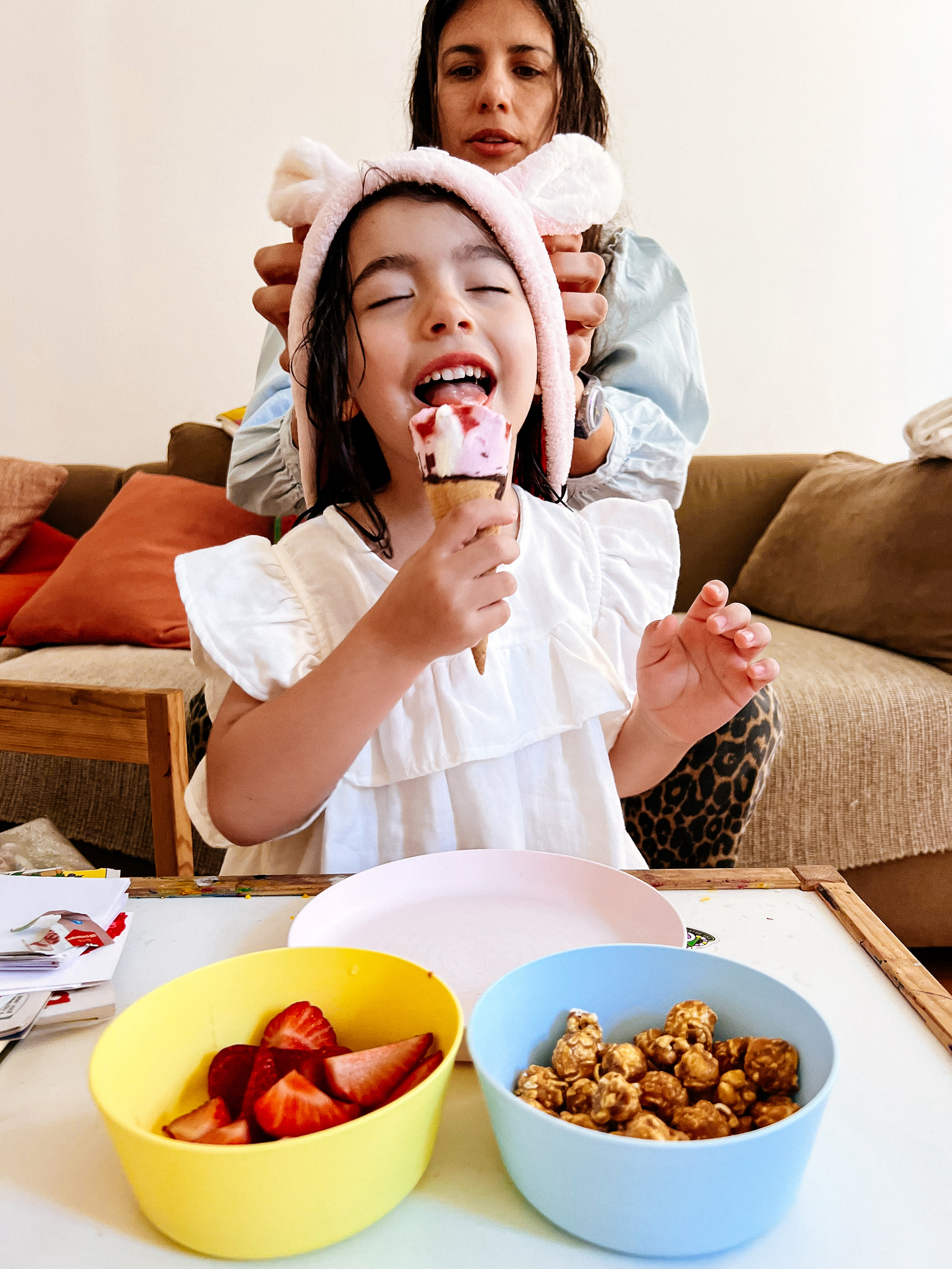 A young girl enjoys an ice cream cone with her eyes closed in delight while an adult woman adjusts a headband with bunny ears on the girl's head. In the foreground, there are two bowls, one with strawberries and the other with caramel-coated popcorn. 